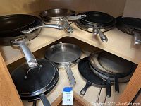 Overview photo showing multiple frying pans and skillets including Calphalon, All-Clad, Wolfgang Puck, and cast iron skillet. Some pans have lids.