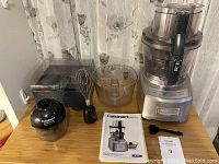 Full set of Cuisinart food processor, smart stick, extra blades, whisk, small chopper, large bowl, small bowl, and instruction manual displayed on wooden table.