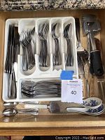 Drawer containing organized stainless steel silverware in plastic tray with knives, forks, spoons, along with a few larger utensils including meat tenderizers and ceramic spoons.