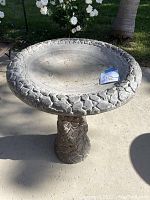 Full view of the gray cement bird bath showing the rock texture on the pedestal and rim, placed on outdoor concrete.