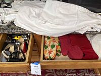 Drawer containing six aprons, two chef hats, and six oven mitts including one floral and red patterned, also Siliconzone trivet