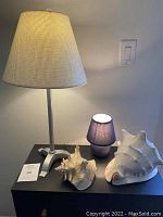Photo showing two large decorative sea shells next to a small blue lamp and a large metal-colored lamp with beige wicker-style shade.