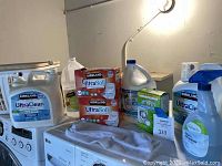 Overview of all laundry supply items displayed on laundry machines including detergent jugs, boxes of dryer sheets, fabric refresher, bleach, and laundry baskets.