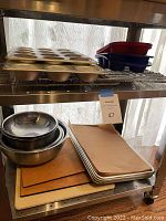Photo shows metal mixing bowls stacked inside each other on a metal shelf. Above them are two metal muffin baking tins, two silicone muffin molds in red and blue, and two wire cooling racks. Below are four metal baking pans stacked, two newer and two used, and a cooking stone at the bottom.