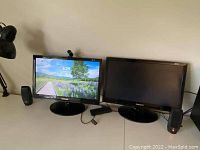 Two Samsung SyncMaster P2250 monitors side-by-side on a white desk. Left monitor shows Windows login screen. Right monitor is off or dark. Each monitor has a black Logitech desktop speaker on either side. Black Microsoft HD webcam attached on top of left monitor. Connectors and cables visible on desk.