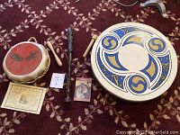 Full view of all three items displayed on a carpeted floor: Pueblo drum with red dragonfly painting, Irish Bodhran drum with blue and gold paint, and the German two-piece recorder along with a small mallet and some paperwork.