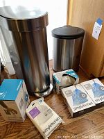 Photo showing the two stainless steel Simplehuman trash cans, one tall 13-gallon and one small round, next to multiple boxes of Simplehuman brand tailor fit trash bags and recycling liners on a wooden floor.