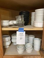 Shelves with white ceramic ramekins, soup bowls, black textured bowls, glass ramekins, and plastic chip bowls organized on two levels in a wooden cabinet