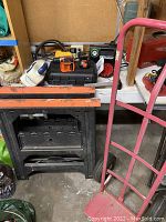 Photo of two black and orange work sawhorse stands, upright red hand truck with wheels, and some small tools on table in background
