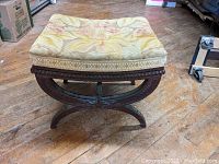 Front angle of the curule stool showing floral needlework upholstery and carved wooden frame.