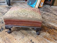 Full view of antique upholstered footstool with patterned tapestry and carved wooden legs on worn wooden floor