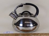 Side angle view of a shiny stainless steel tea kettle with black handle and lid knob, showing rounded body and spout with lever for whistle cover.