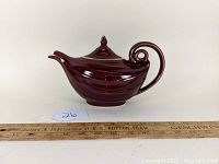 Side angle view showing dark maroon ceramic teapot with spiral handle, spout and lid on ruler for size reference.