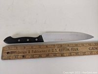 Chef's knife placed next to ruler showing ~8 inch blade length, black handle with rivets and polished stainless steel blade