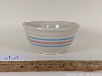 Side view showing the pink and blue bands on the ceramic bowl with a flared lip, placed next to a wooden ruler for size reference.