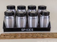 Side view showing 8 metal jars in black metal spice rack labeled SPICES with ruler for scale.