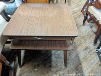 Top and front view of the mid-century style end table showing the laminate wood grain top, drawer with metal handle, and open shelf below.