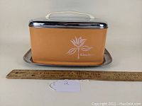 Side view of the rectangular beige-orange cake carrier with chrome lid, white floral design on the side, and a ruler for size reference.