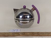 Front-left angle view showing the spherical metal body, purple handle and lid knob, and spout. Ruler for scale.
