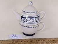 Side angle of the stacked teapot and teacup showing crackled white ceramic with dark blue decorative band and floral motifs