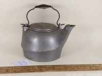 Side view of 19th-century cast aluminum tea kettle with coiled wire handle, showing kettle body, spout, and lid with embossed signature marks.