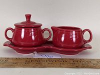 Side view of red Fiestaware creamer, sugar bowl with lid, and tray beside a ruler for scale.