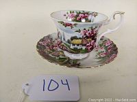 Porcelain teacup and saucer showing front view with pink rose floral decorations and cottage scene, gold rim and handle accents.