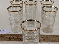 Set of six clear glass tumblers with embossed ring patterns, gold rim and base trim, each approximately 5 inches tall, arranged showing sides and fronts.