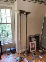 Pair of gold-colored floor lamps standing near window and wall with framed pictures on the floor beside.