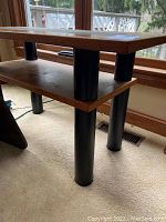 Angled view of the table showing the teak frame, black cylindrical legs, and lower shelf.