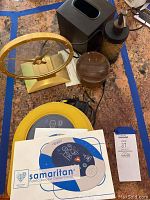 All lot items displayed on countertop: yellow defibrillator case with manual and adapter, gold desk clock, black tissue box cover, marbled soap dispenser, wooden container.