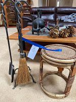 Photo of iron fireplace tools with twisted decorative handles including broom, shovel, and poker, placed next to a round wicker table with woven top and wooden frame.