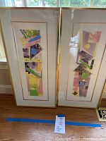Two tall, narrow framed abstract art prints with pastel geometric shapes and patterns, leaning against a wall on a wooden floor.