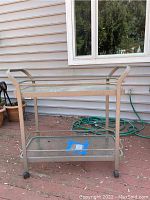 Front-right angled view of a metal garden cart with frosted glass shelves and casters on an outdoor deck, showing overall structure and condition.