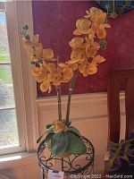 Yellow faux orchid with multiple delicate flowers and lifelike green leaves displayed on black iron plant stand.