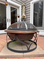 Front view of the portable fire pit sitting on four cement tiles with metal mesh dome cover in place on the stand with decorative hammering on the copper rim and sturdy black metal legs.