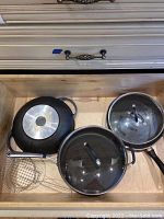 Drawer with a large black non-stick skillet, a large metal pot with glass lid, a medium saucepan with glass lid, and wire basket skimmer.