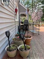 Photo showing five beige outdoor planters on a wooden deck, some with plants growing inside. Seven black patterned solar stake lights placed in the planters. Displayed next to the side of a house and deck railing in background.