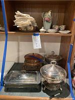 Shelf showing silver plate serving dishes, copper chafing dish, cloth napkins, china tea set components, decorative vase