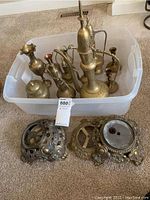 Overview of multiple brass items inside a plastic tote, including pitchers, candlestick holders with wooden handles, and a small brass vase, with four ornate metal bases placed outside the tote on the carpet.