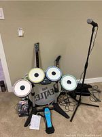 Electronic Ludwig drum kit setup with drum pads in red, yellow, blue, and green, bass drum pad with Beatles logo, black guitar controller, microphone on stand, and several Xbox 360 game cases on floor.