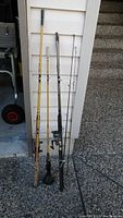 Three fishing rods leaning against a white exterior wall near a garage. Includes one fly rod with green reel, one black and yellow spinning rod with reel, and one yellow bamboo or fiberglass rod.