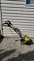 Sunjoe 16-inch electric tiller cultivator with black handle, yellow-green body, and wheels for soil cultivation, shown standing upright outdoors on gravel with manual placed on top.
