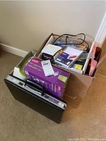 Overview photo showing cardboard box filled with assorted items including a purple boxed desk lamp, papers, and other supplies, with a black briefcase in front on carpet.