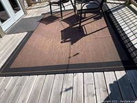 Large brown outdoor rug with solid brown center and black border, placed on wooden deck with outdoor chairs.