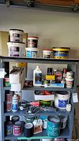 Shelf with stacked tubs of dust control compound, bonded topping, paint thinner, containers of paints, paint cans, brushes, rollers, and other painting and maintenance containers.