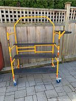 Full view of yellow Metaltech rolling scaffolding with three black platforms and locking casters against wood fence.