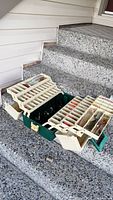 Opened green Plano tackle box showing multiple beige fold-out compartments filled with fishing tackle items.