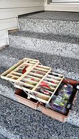Tackle box opened on stairs showing upper compartments filled with assorted fishing lures and hooks.