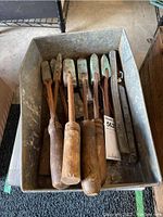 Metal container holding six vintage hand tools with wooden handles and metal blades showing rust and wear, plus several metal bars and hardware pieces.